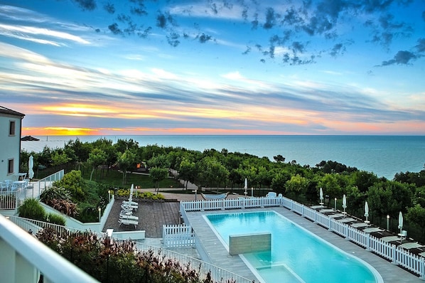 Ein Schwimmbecken mit weißem Zaun, Liegestühlen und Blick auf das Meer bei Sonnenuntergang.
