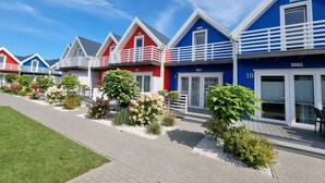 Una hilera de casas de playa coloridas con barandillas blancas y números en las puertas, rodeadas de jardines cuidados y un camino pavimentado.