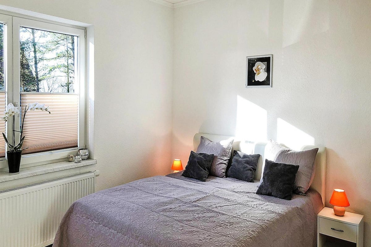 Ein Schlafzimmer mit Bett, einem Fenster mit Jalousien, einem Nachttisch mit Lampe und einem Bild an der Wand.