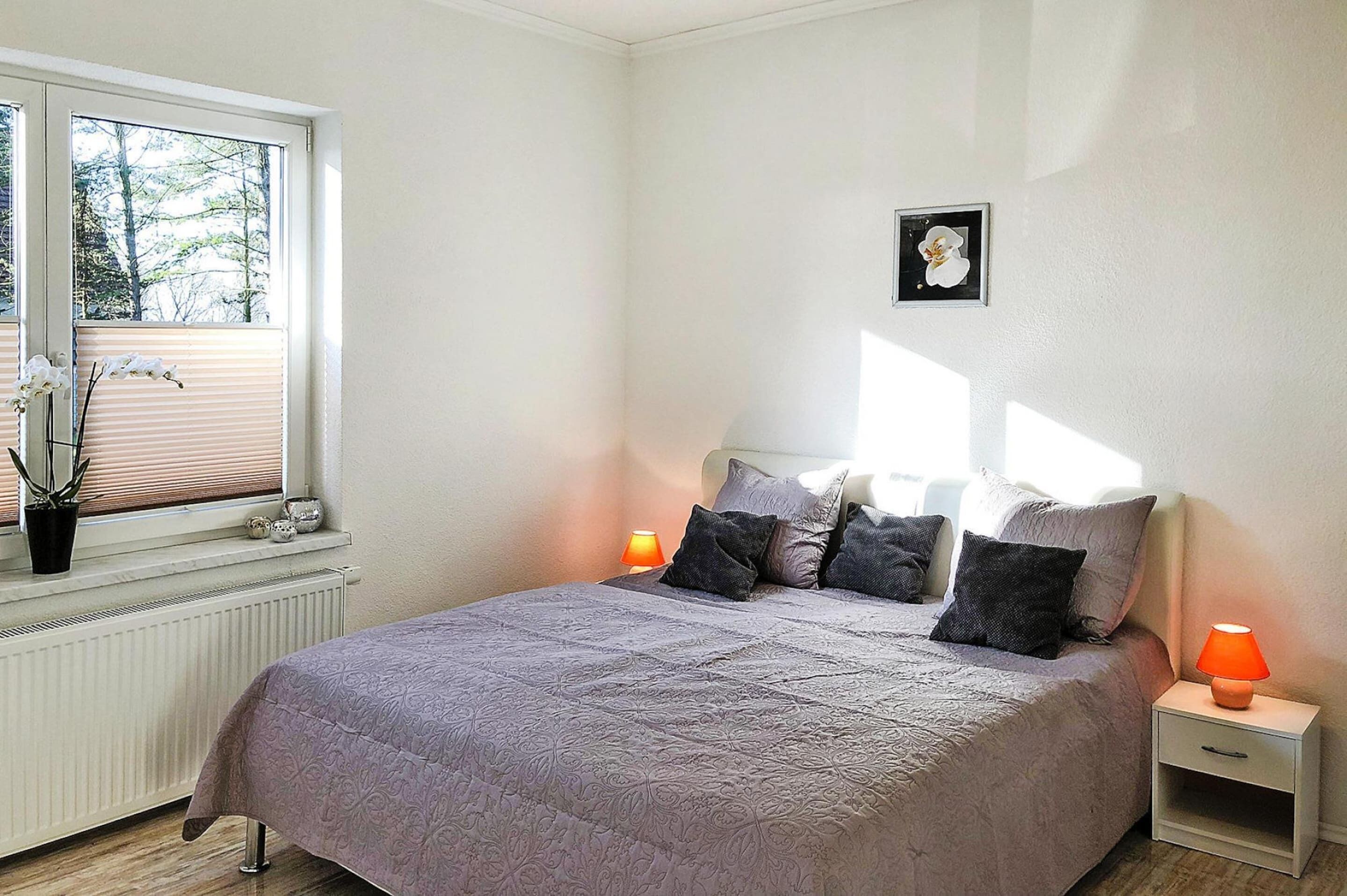 A bedroom with a bed, two bedside tables, a window with blinds, a radiator, and a potted plant.