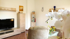 Un soggiorno con una TV a schermo piatto, una poltrona beige, un tavolino con un vaso di fiori e una libreria.