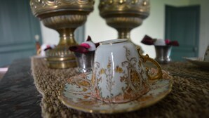 A white teacup with gold patterns on a matching saucer, placed on a woven mat.