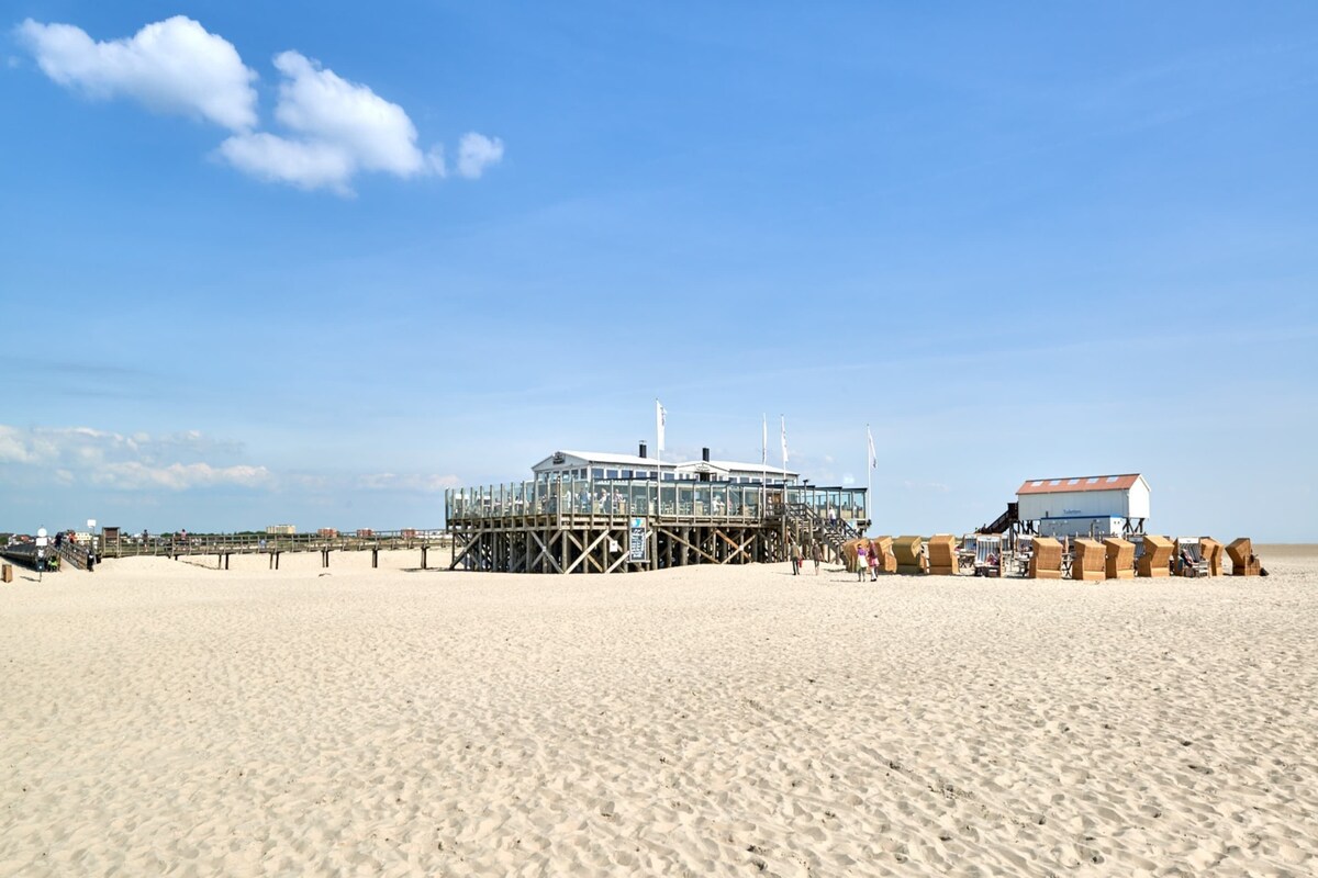 A beach with a wooden pier leading to a glass-enclosed building, likely a restaurant or bar, and a smaller building with a red roof.