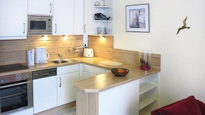 A compact kitchen with white cabinets, a microwave, a toaster, and a sink. There is a small island with a bowl and a vase. A framed picture hangs on the wall, and a red towel is on the floor. - Apartment in Cuxhaven With Community Pool (Cuxhaven)