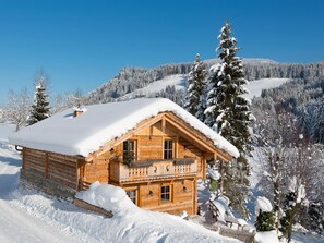 Une cabane en rondins de bois dotée d’un balcon, entourée d’arbres couverts de neige et avec une chaîne de montagnes en arrière-plan.