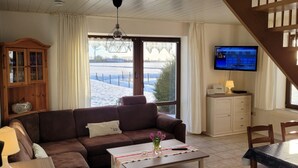 A cozy living room with a brown sofa, a coffee table with a vase of flowers, a television on the wall, and a view of a snowy landscape through the window.
