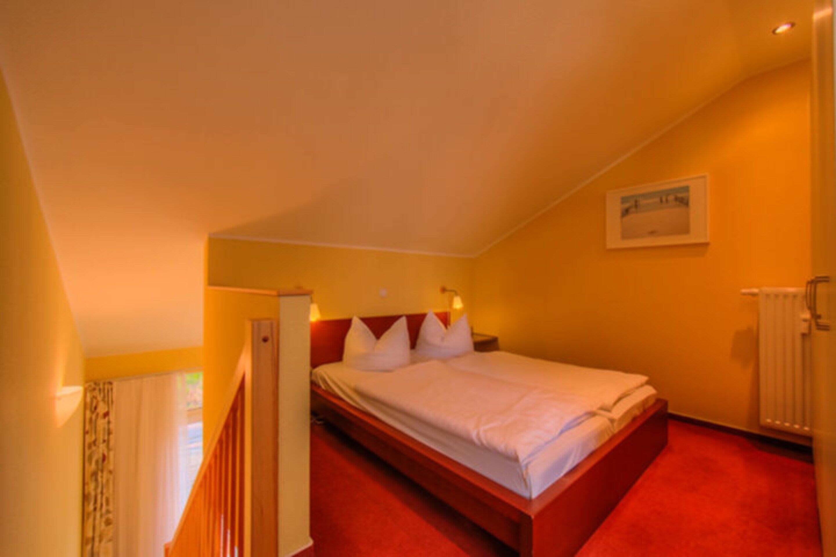 Camera d'albergo con un letto singolo, una testiera in legno, un tappeto rosso, una porta bianca e un piccolo quadro appeso al muro.