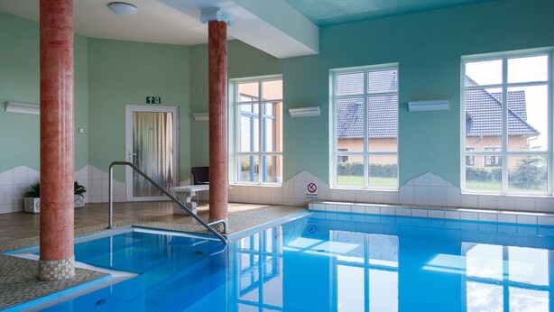 Una piscina interior con paredes de vidrio transparente, suelo de baldosas y vista a una casa a través de amplias ventanas.