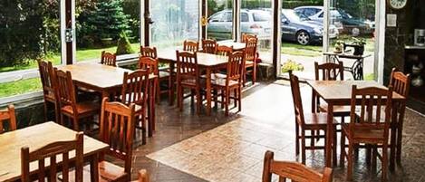 Un espace de restauration extérieur abrité, doté de tables et de chaises en bois, de grandes fenêtres et offrant une vue sur la verdure environnante.