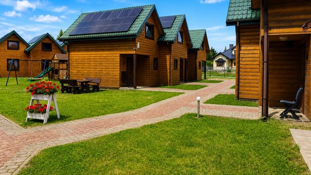Wooden cabins with green roofs and solar panels, a brick pathway, and a flower display.