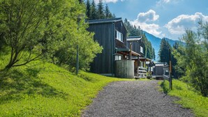 A wooden cabin with a gravel path leading to it, surrounded by lush greenery and mountains.