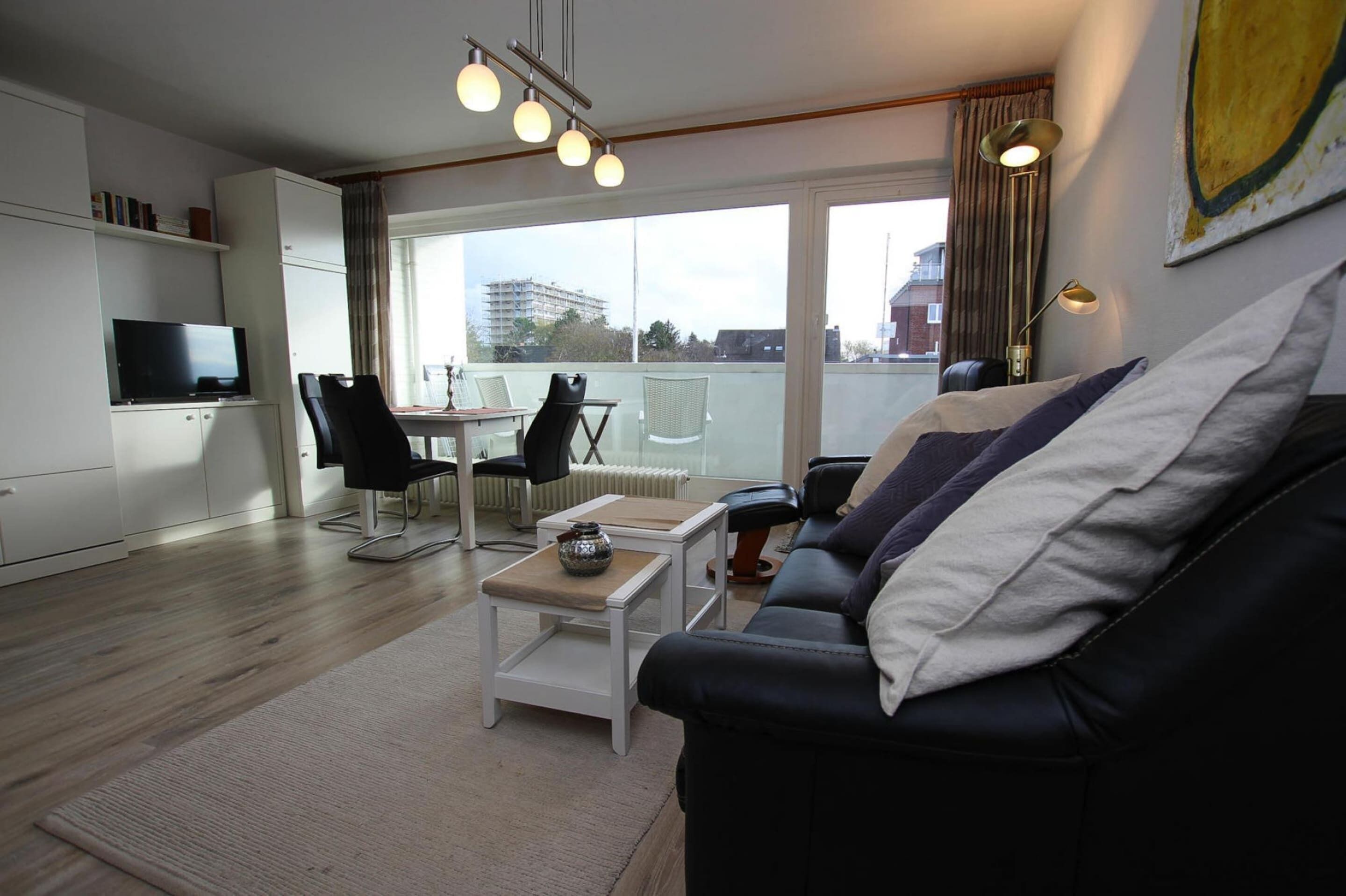 Una sala de estar moderna con un sofá de cuero negro, una mesa de centro blanca y un televisor de pantalla plana. Una ventana grande ofrece vistas de la ciudad.
