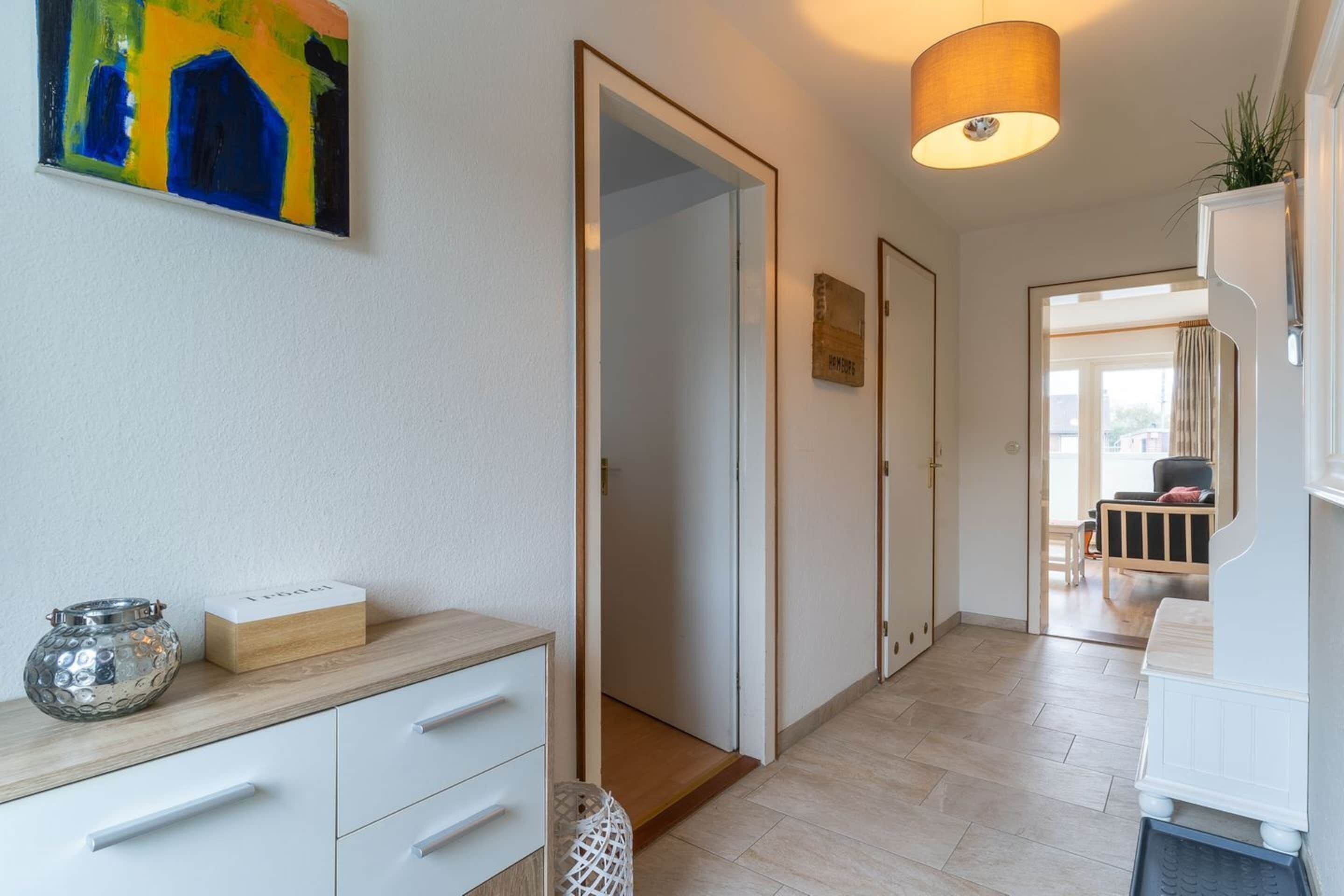 A hallway with a wooden floor, white walls, and a door leading to a room. There is a small table with a vase and a box on it, and a painting on the wall.