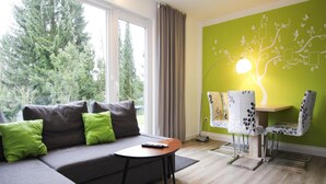 A modern living room with a grey sectional sofa, a small round table, and a green wall with a tree design. - Ferienwohnung Hexenzauber I und II in Braunlage (Braunlage)
