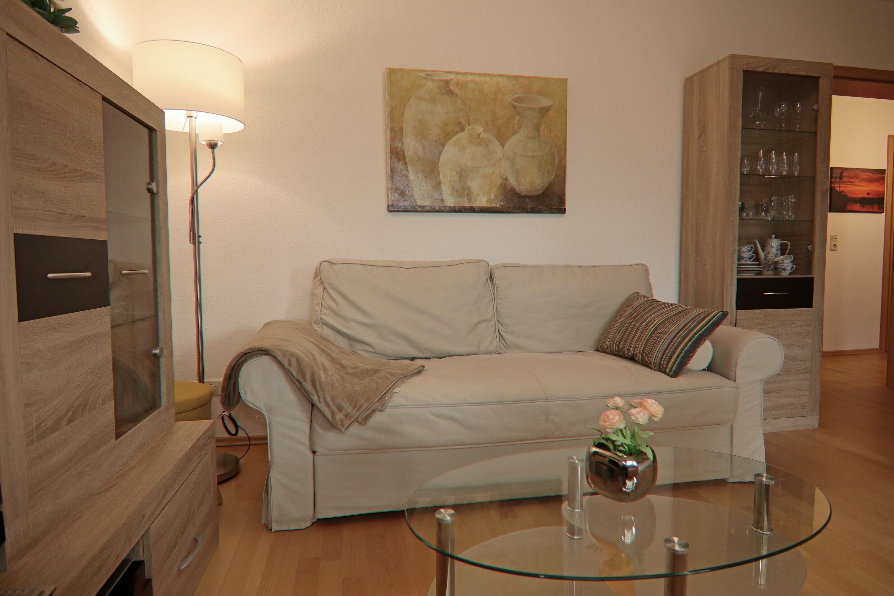Una sala de estar con un sofá color beige, una mesa de centro de cristal, una lámpara y un cuadro en la pared.