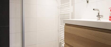A modern bathroom with a glass shower enclosure, a white tiled wall, a wooden vanity with a sink, and a mirror above the mirror.
