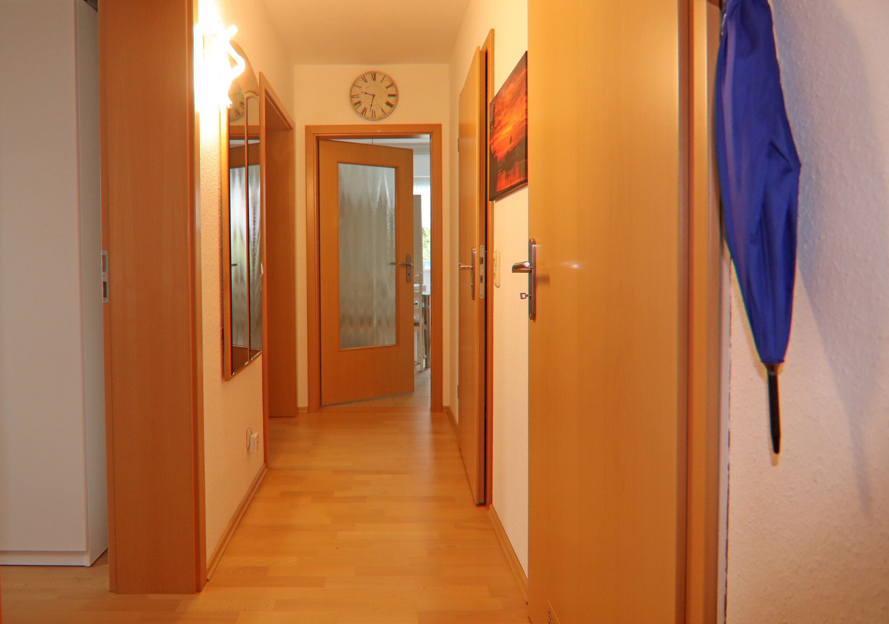Een hal met houten vloer, een klok aan de muur en een deur aan het einde.