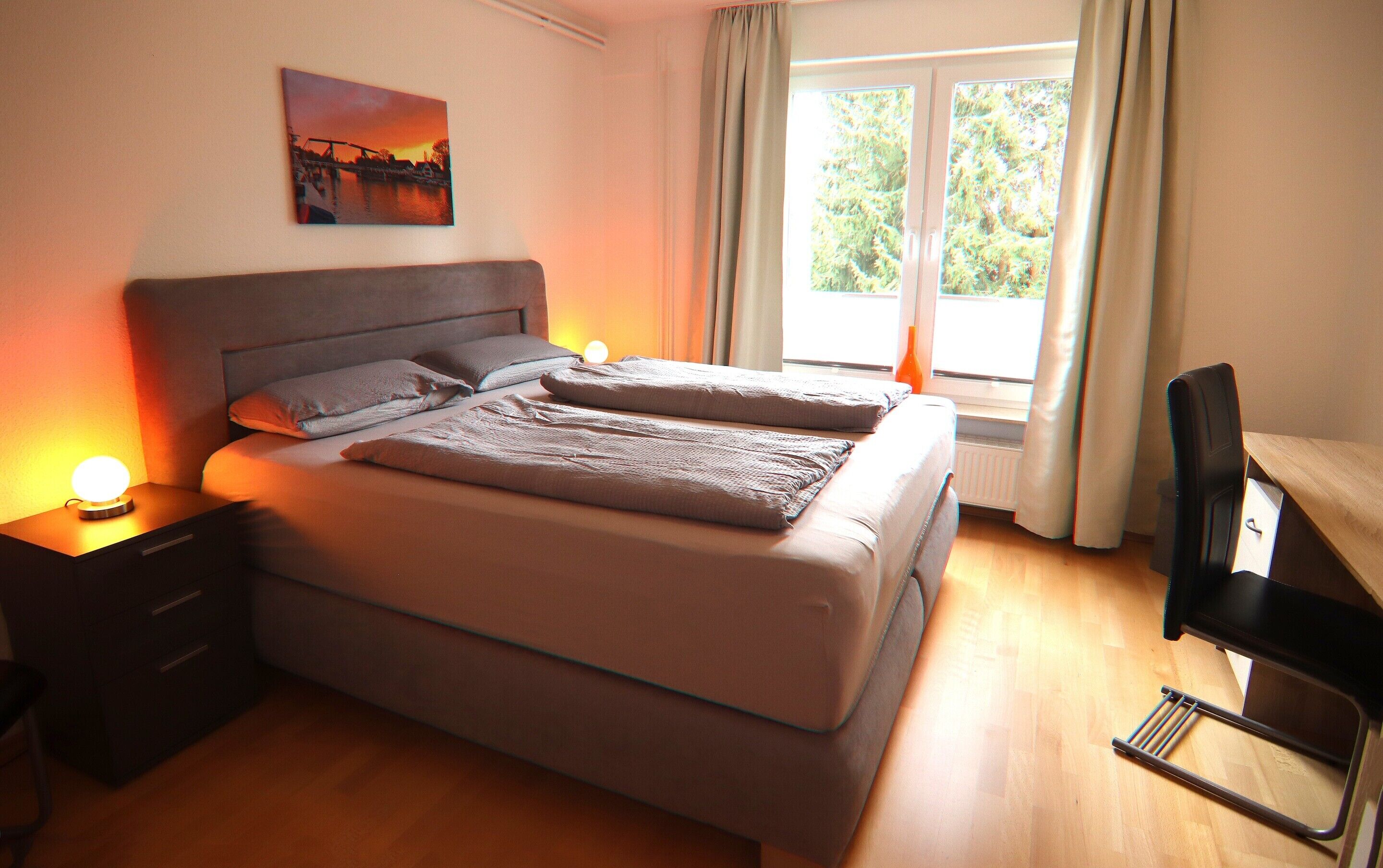 A double bed with a headboard, a bedside table with a lamp, a chair, and a window with curtains.