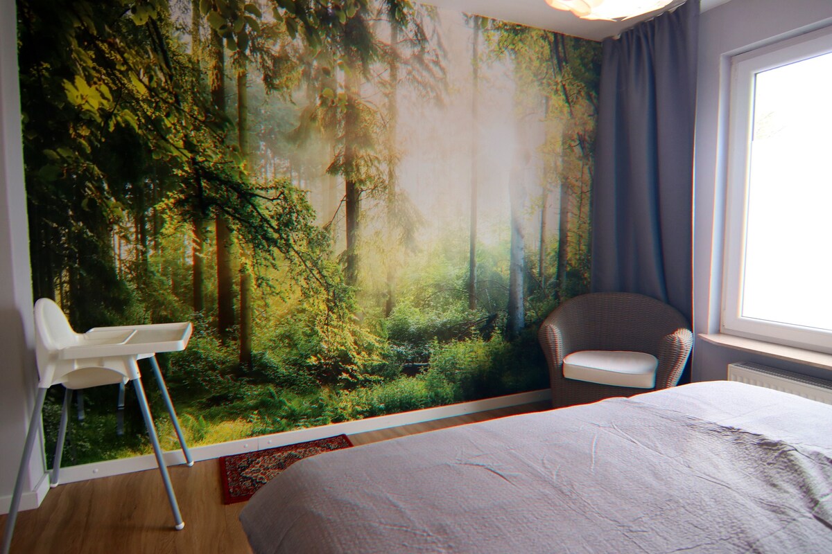 A bedroom with a large forest mural, a bed, a chair, and a window with curtains.