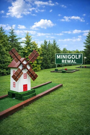 A model windmill with a red door, a sign reading 'MINIGOLF REWAL,' and a green field with a clear sky.