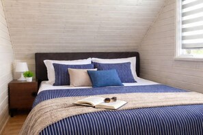 Een slaapkamer met schuine wand, een donker hoofdbord, een bed met gestreepte lakens, een nachtkastje met een lamp, en een raam met blinds.