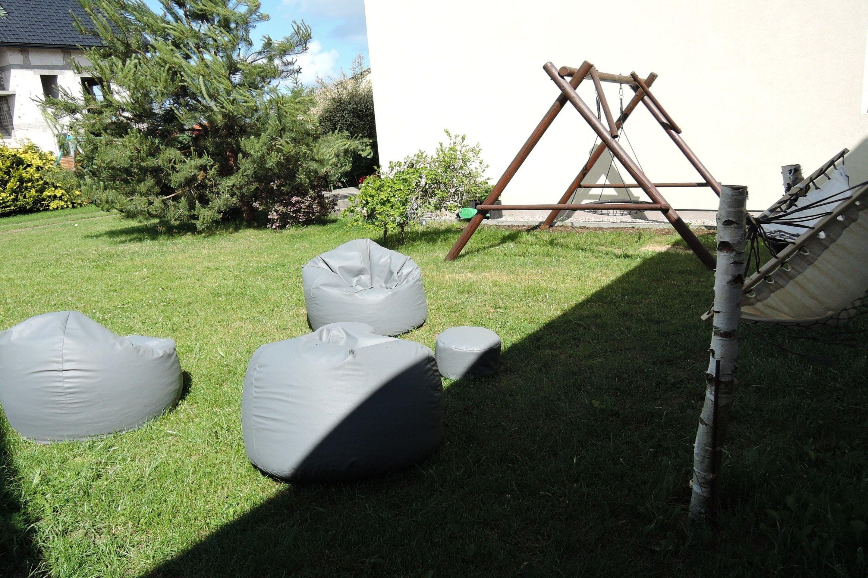 A grassy outdoor area with three gray bean bags, a wooden swing set, and a white wall in the background.