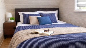 A bedroom with a sloped ceiling, a bed with striped bedding, a bedside lamp, and a window with blinds. - Holiday Home in a Holiday Park Rewal at 600 m From the sea (Rewal)