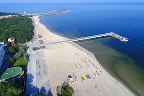 Luchtbeeld van een strand met een lange pier die in het water uitloopt, een zandstrand met parasols en een gebouw met een groen dak.