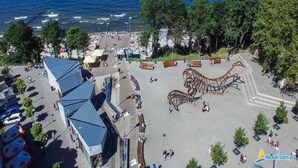Luchtbeeld van een kustpromenade met een grote metalen sculptuur, een parkeerplaats en uitzicht op zee.
