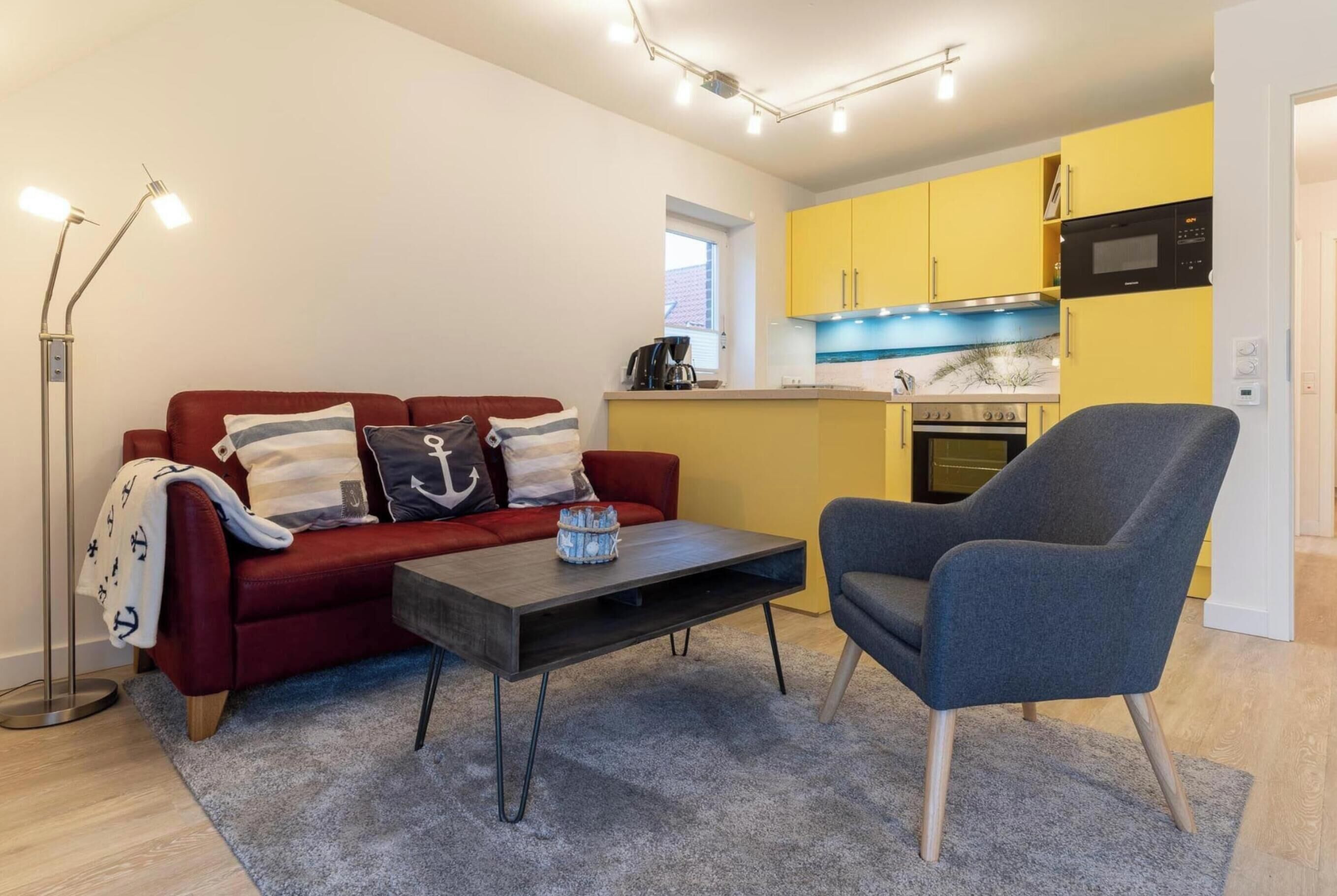 Ein kompakter Wohnbereich mit rotem Sofa, grauem Sessel, Couchtisch, Stehlampe und einer Küchenzeile mit gelben Schränken und modernen Geräten.
