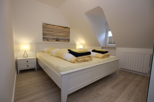 Ein Schlafzimmer mit einem weißen Bett, einem Nachttisch, einem Bild an der Wand und einer schrägen Decke.
