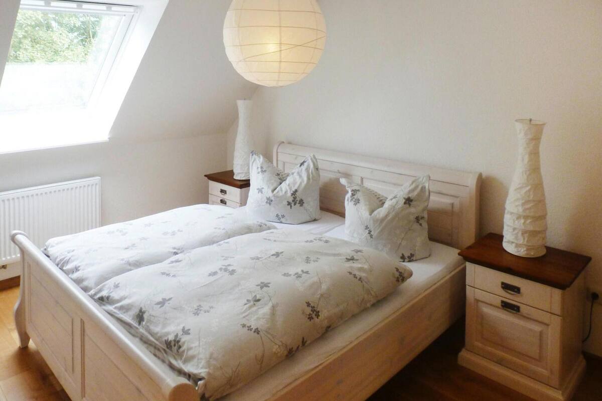 Un dormitorio con una cama de madera, ropa de cama blanca con motivos florales, una mesita de noche blanca con un florero y una ventana con vistas a la vegetación.