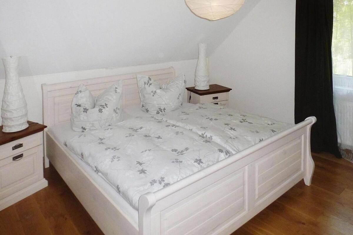 Una cama blanca con ropa de cama floral, una mesita de noche con un jarrón, y una lámpara colgante.