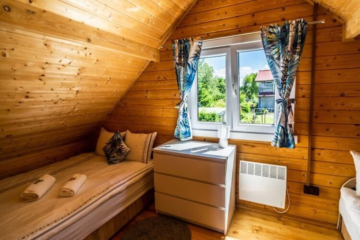 Una habitación pequeña y acogedora con paredes de madera, una cama individual, una mesita de noche, un radiador y una ventana con cortinas.