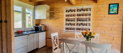 A wooden interior with a kitchen area, white cabinets, a dining table with chairs, and a wall-mounted shelf with potted plants.