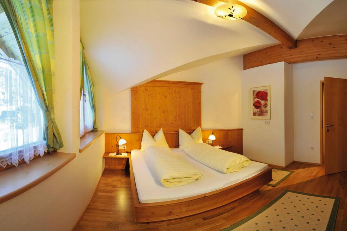 Ein Schlafzimmer mit einem Holzkopfende, zwei Nachttischen mit Lampen, einem großen Bett mit ordentlich zusammengefalteter Bettwäsche, einem Fenster mit Vorhängen und einer Holztür.