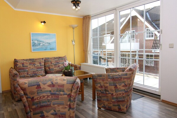 A living room with patterned sofas, a table with a vase of flowers, a floor lamp, and a large window with a view of a house. - Appartementhaus Pinamar, Scharbeutz (Scharbeutz)