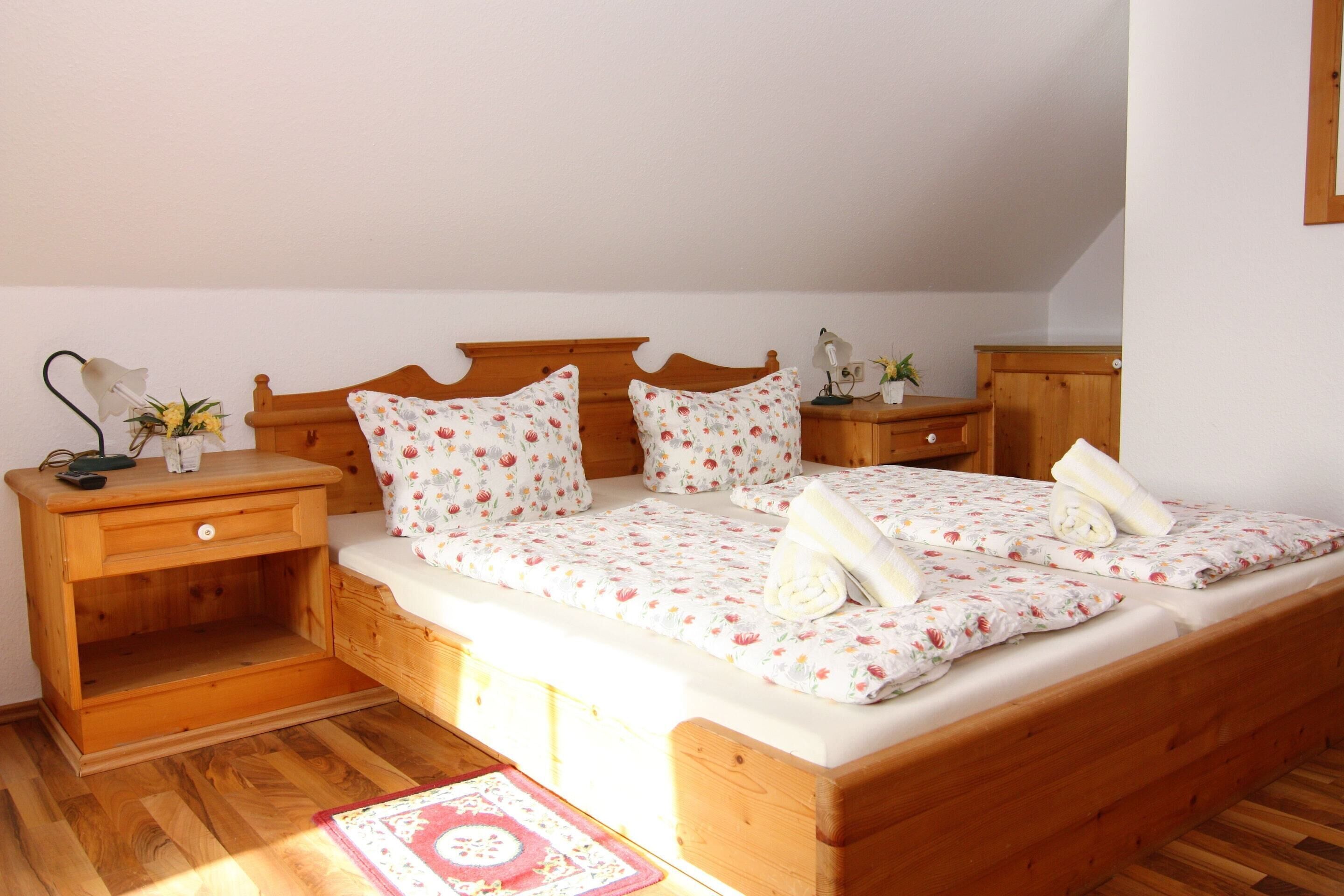 A wooden bed with floral bedding, a nightstand with a lamp, and a small table with a plant.