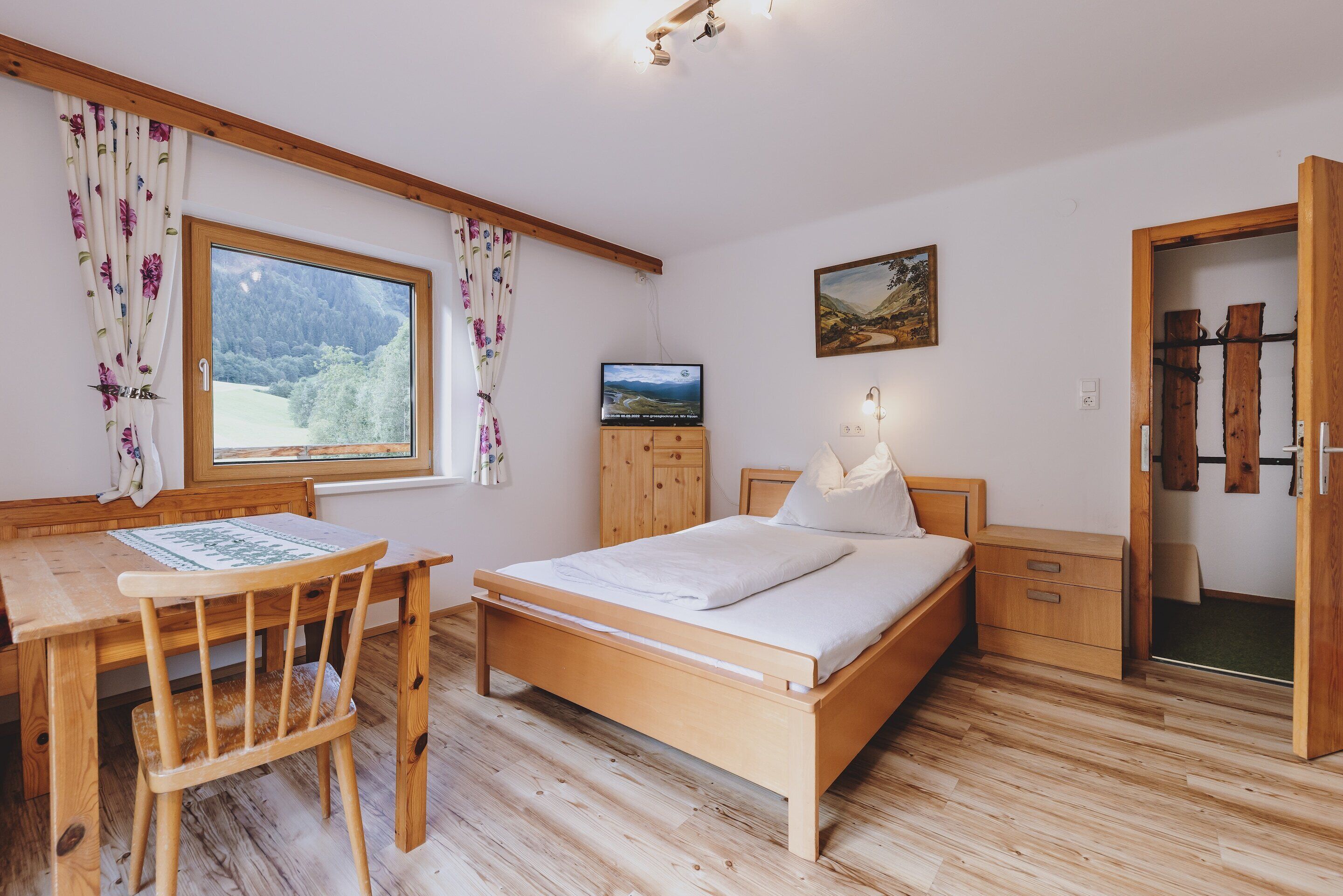 Een slaapkamer met een houten bed, een bureau en een stoel. Er is een raam met gordijnen, een schilderij aan de muur en een houten garderobe.