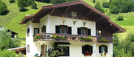 Un chalet de dos pisos con balcón, rodeado de exuberante vegetación y montañas.