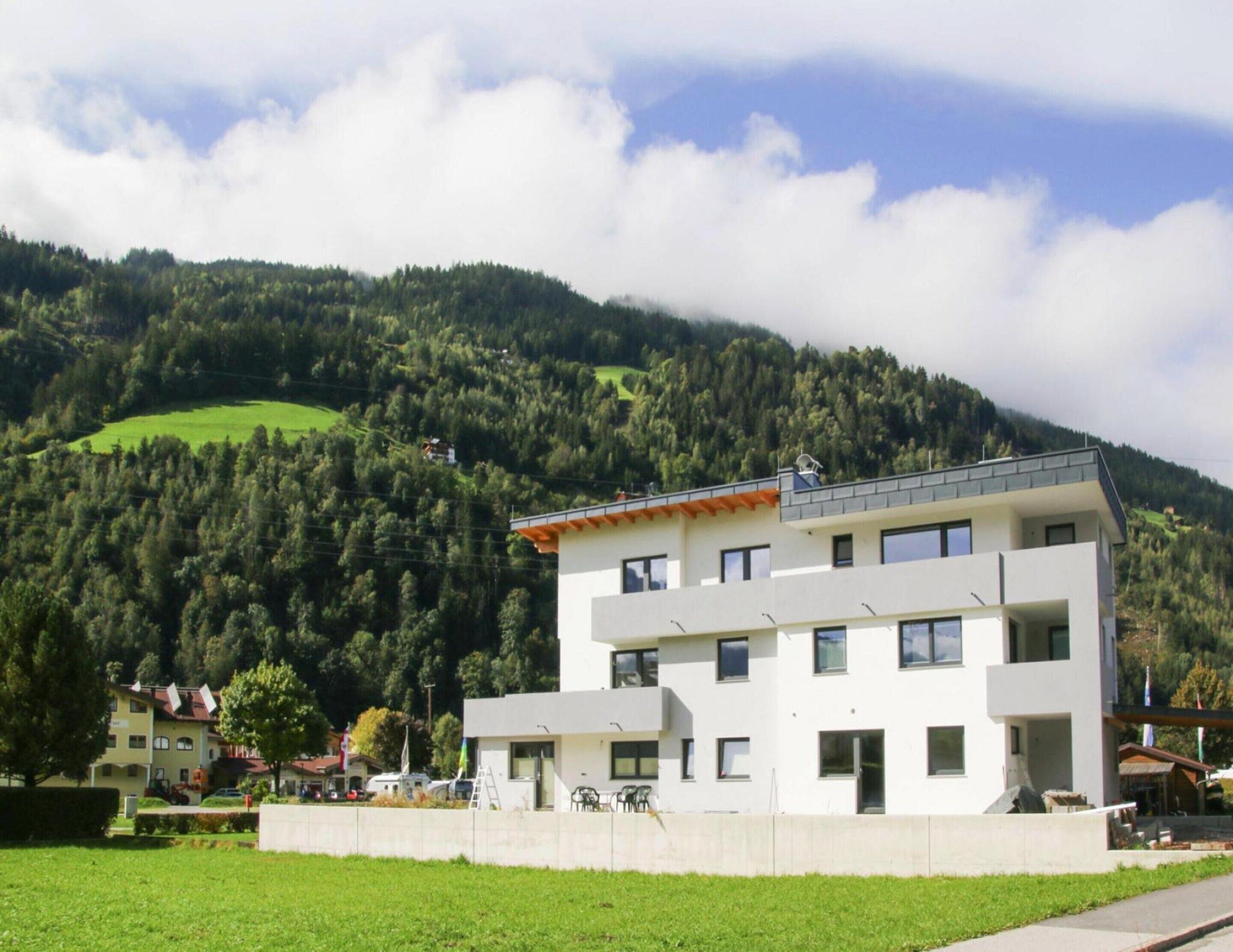 A modern two-story building with a flat roof and large windows, surrounded by a well-maintained lawn and a mountainous backdrop.