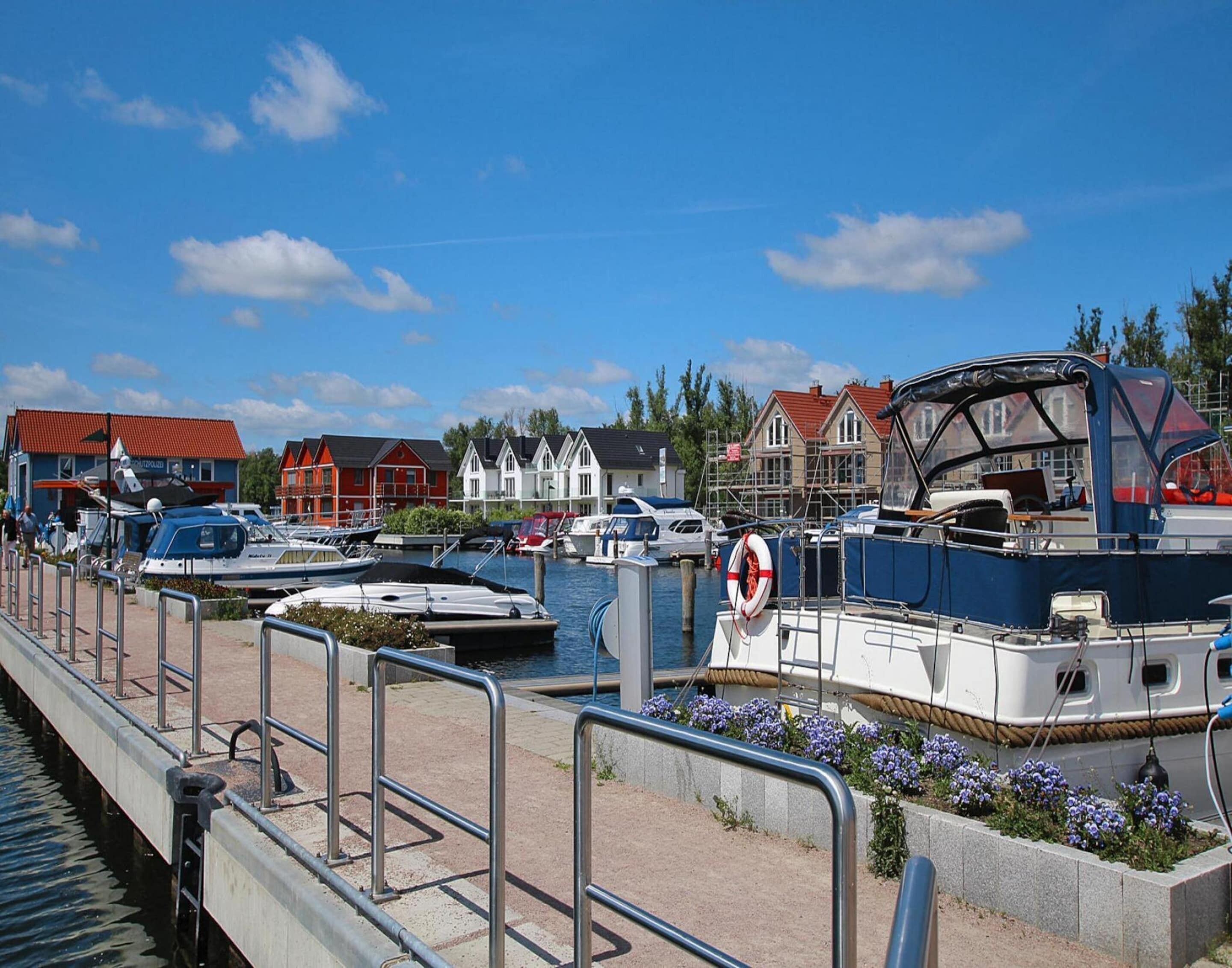 A marina with boats docked, a pedestrian walkway, and buildings with red roofs in the background.