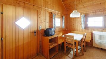 Eine Holzhütte mit Esstisch und Stühlen, einem Fernseher auf einem Regal und einem Fenster mit Vorhängen.