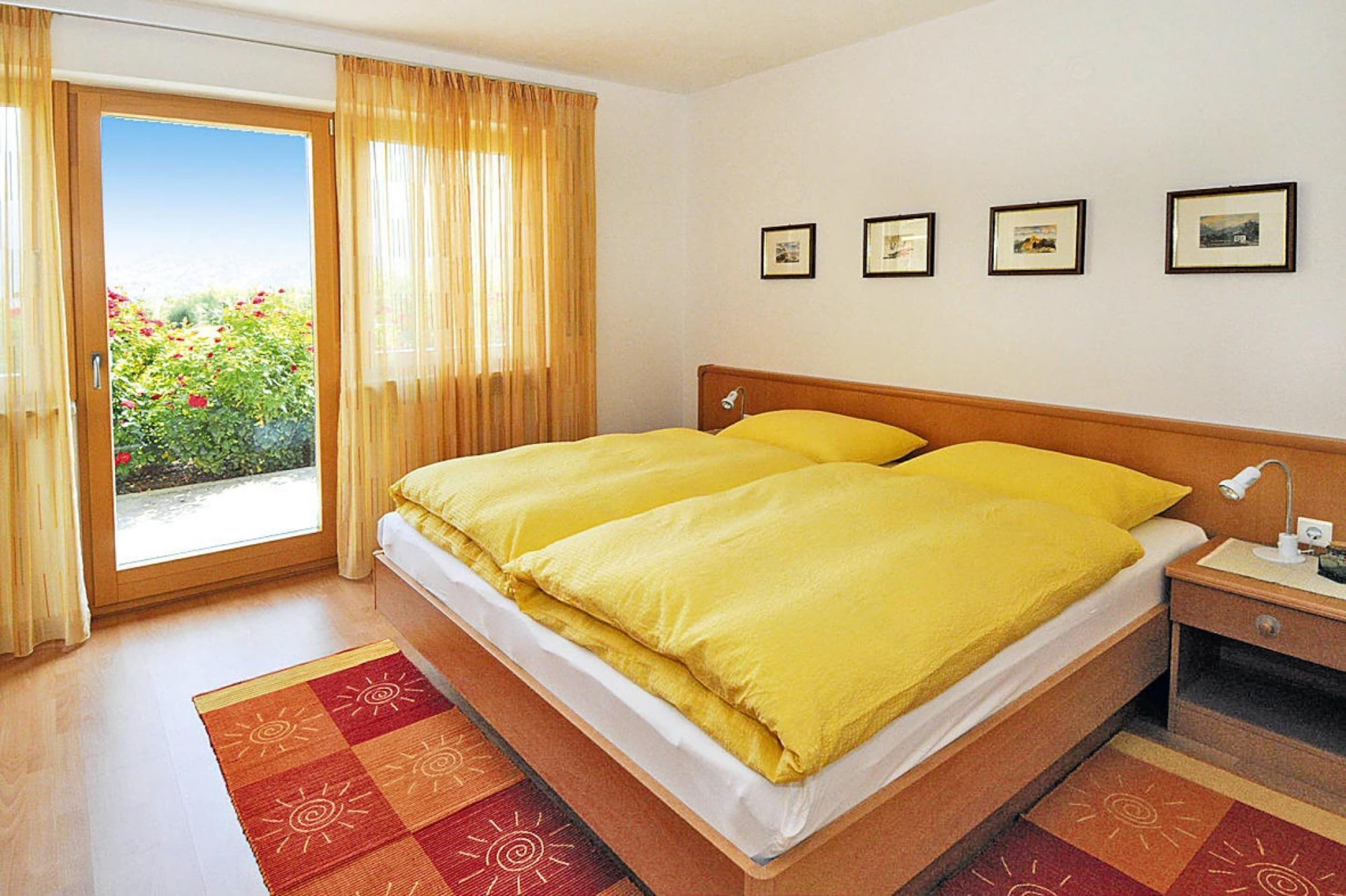 A hotel room with a large bed, a nightstand, a window with curtains, and a view of greenery outside.
