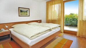 A double bed with a wooden headboard, a nightstand with a lamp, a framed picture on the wall, and a view of greenery through a window with curtains. - Apartment in Schlanders With Terrace (Silandro)