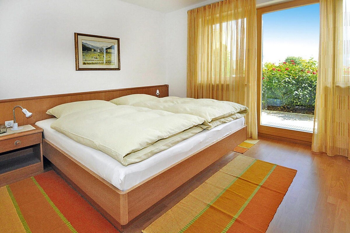 A double bed with a wooden headboard, a nightstand with a lamp, a framed picture on the wall, and a view of greenery through a window with curtains.