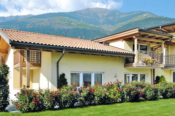 Une maison à deux étages dotée d’un balcon, entourée d’un jardin bien entretenu où poussent des fleurs rouges.