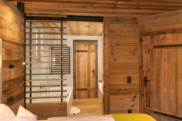 A wooden cabin interior with a bed, a bathroom with a shower, and a door leading to another room.