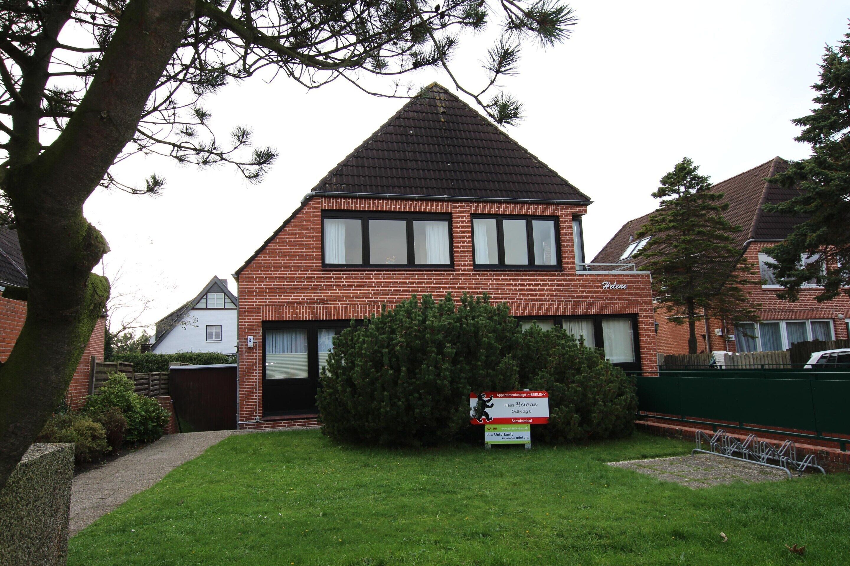 A two-story brick house with a triangular roof, a front yard with a small bush, and a signboard in front.
