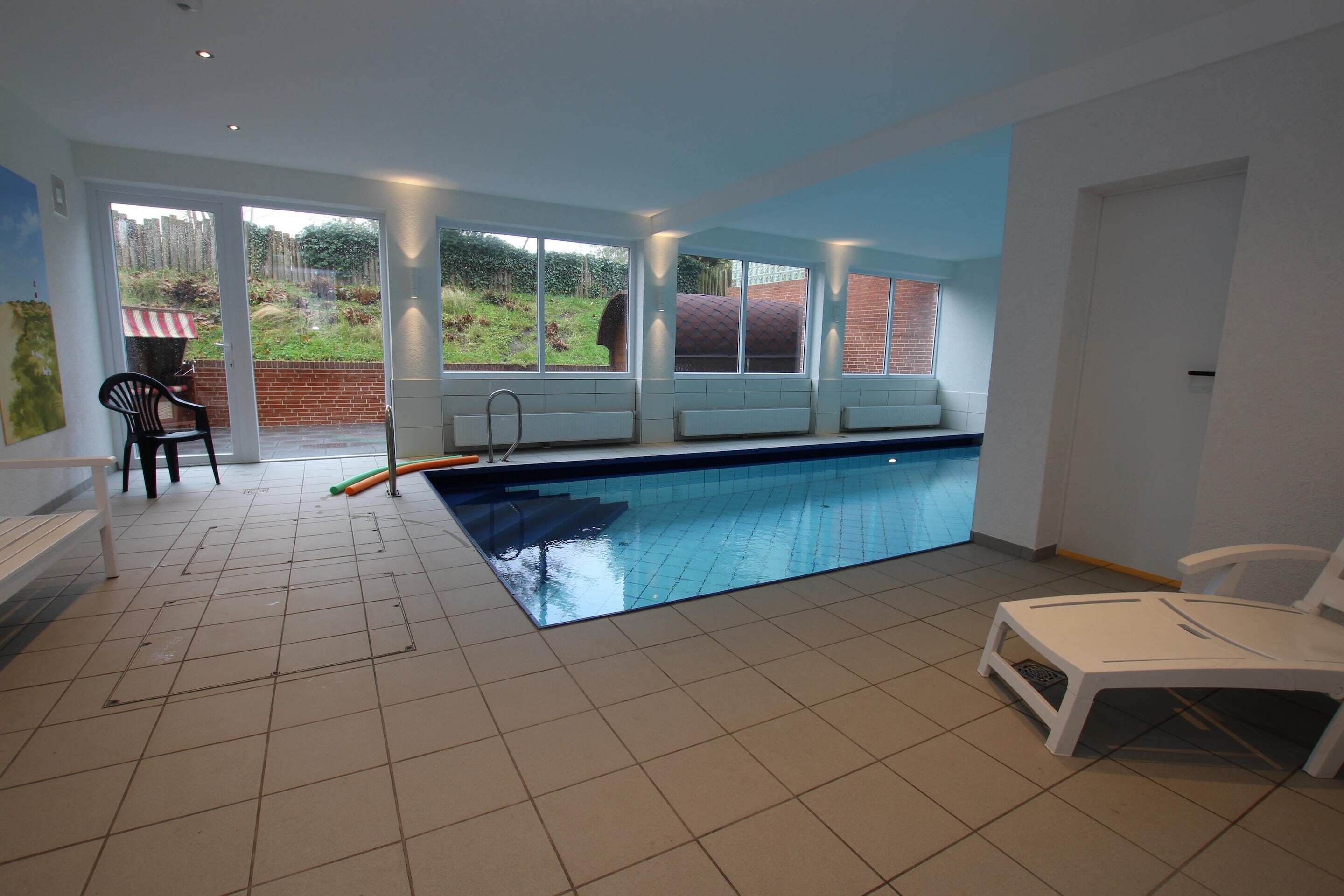 An indoor swimming pool with a view of a brick wall and greenery outside. There are white chairs and a bench near the pool.